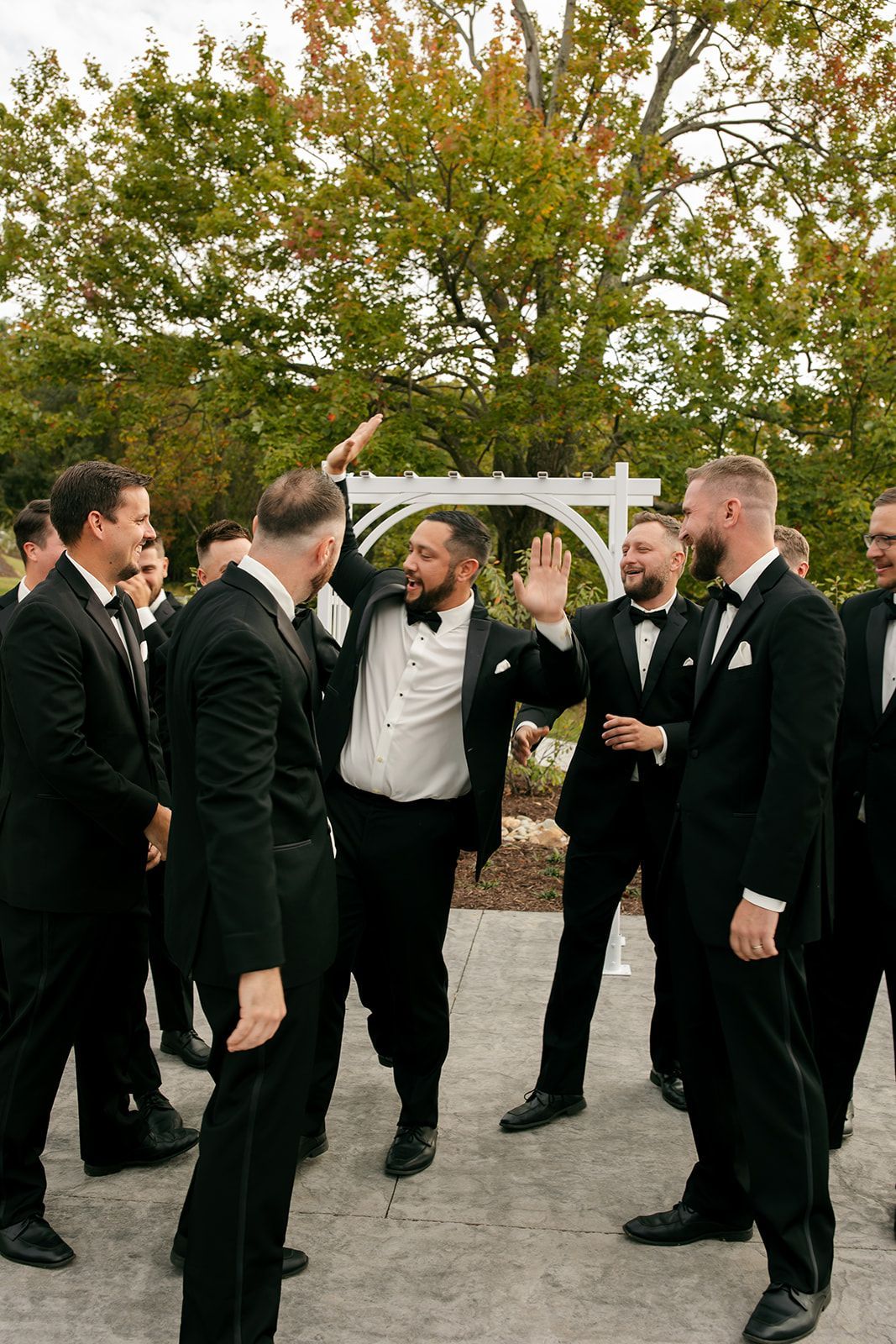 Men in black suits celebrating outdoors under a white archway in a garden.