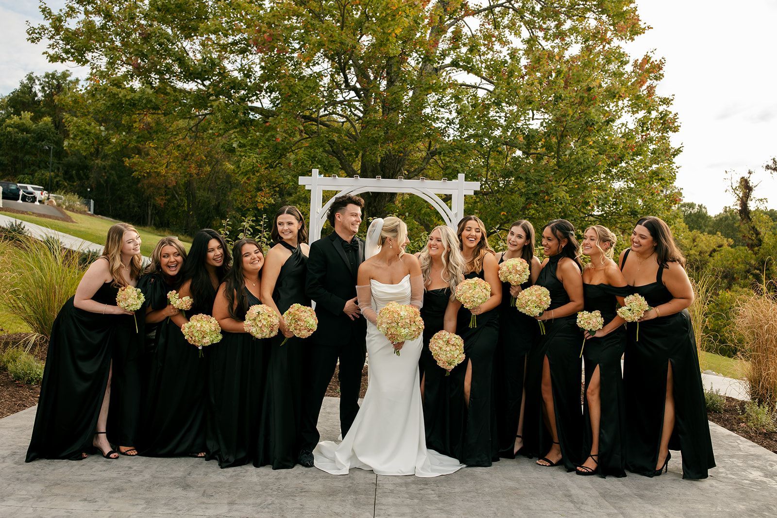 Bride and bridesmaids pose outdoors in black dresses and white gown under a wooden arch, holding bouquets.
