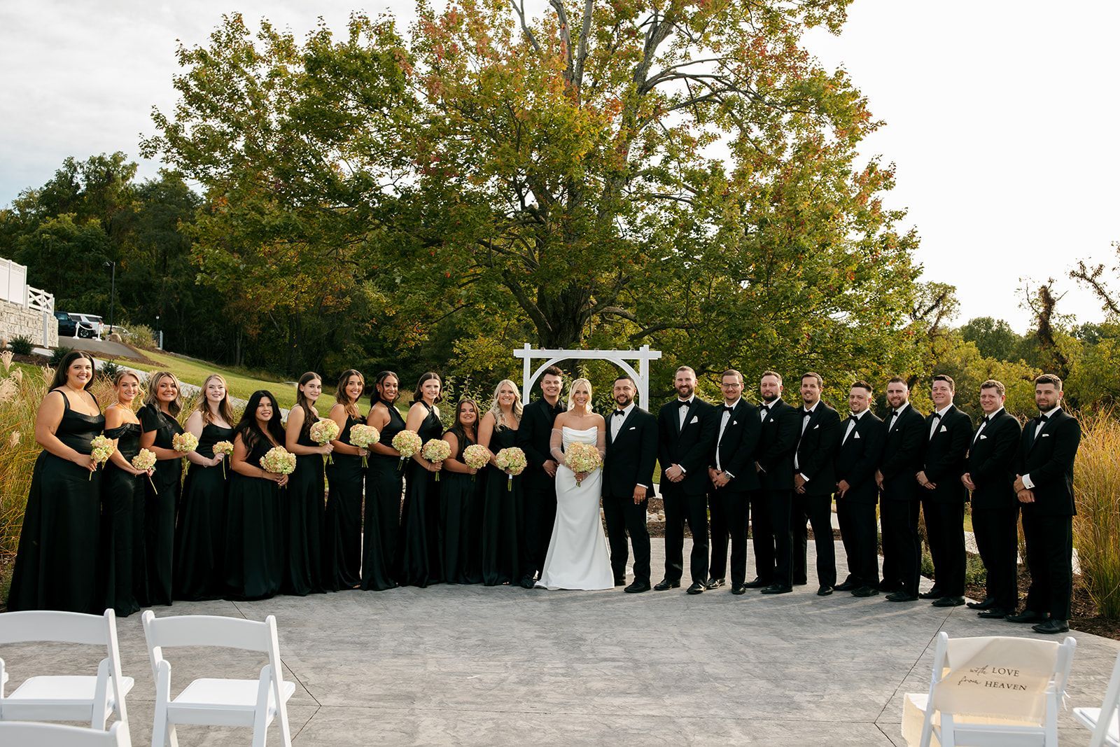 Wedding party posing outdoors under a white arch, with bridesmaids in black dresses and groomsmen in black suits.