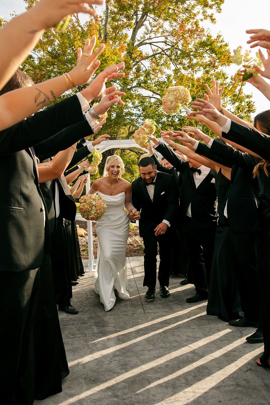Bride and groom walking through an arch of raised hands outdoors under autumn trees