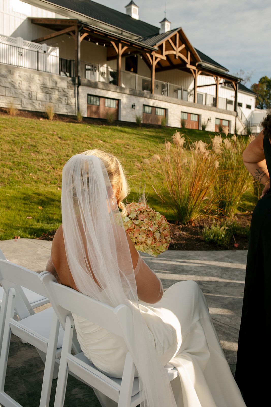 Bride in white veil seated outdoors at a wedding venue, facing a hillside lodge.