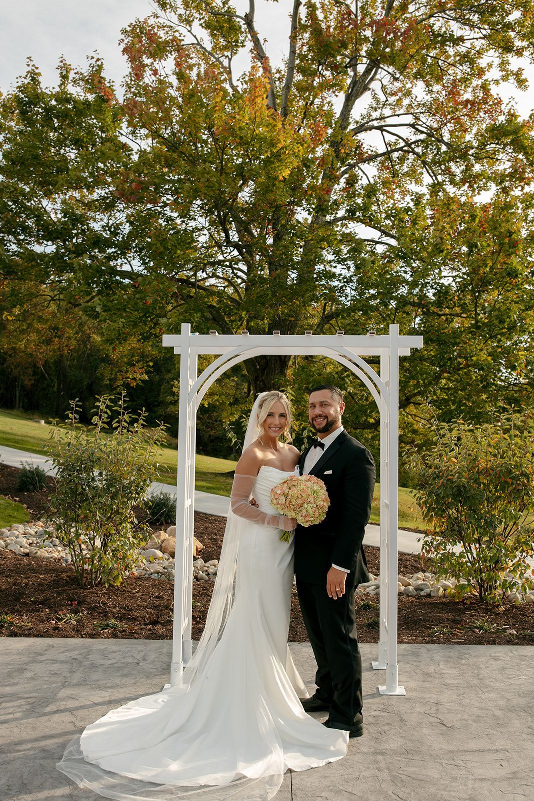 Bride and groom standing under a white arch outdoors, holding a bouquet in front of autumn trees