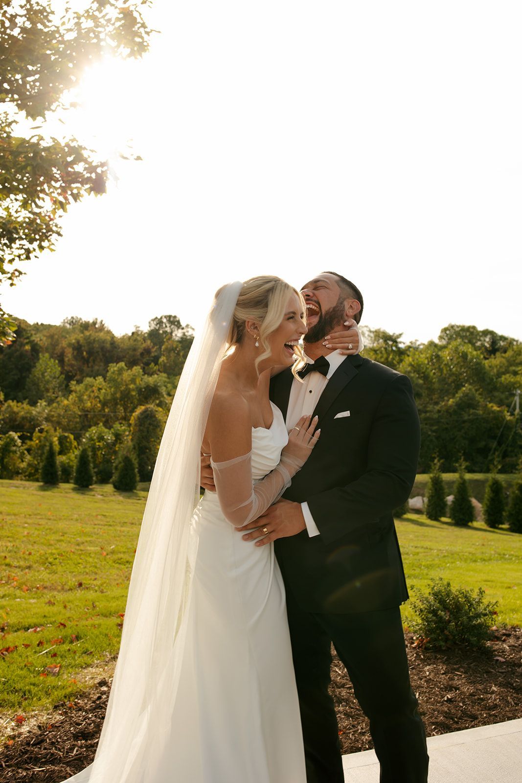 Bride and groom in white dress and black tuxedo embracing outdoors in a sunny garden
