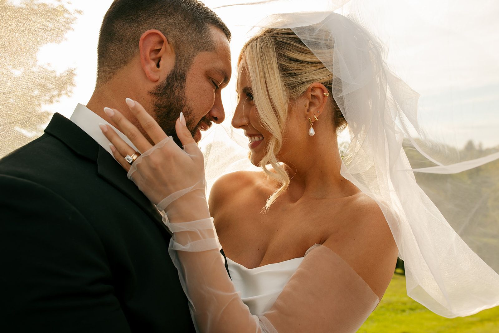Bride and groom embracing outdoors in warm sunlight, smiling beneath the bride’s veil
