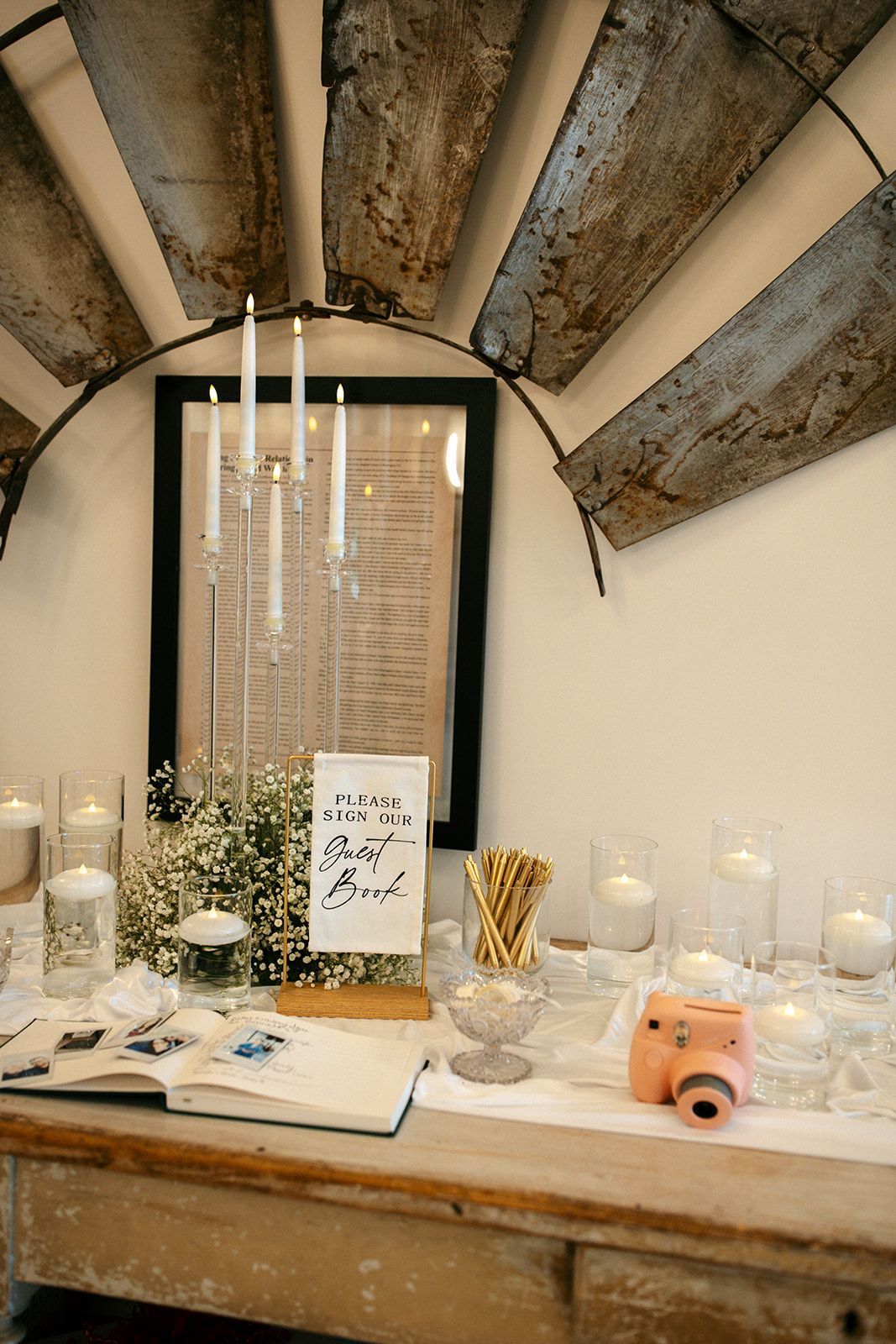 Decorative table display with candles, flowers, books, and a pink instant camera under a rustic fan-shaped wall