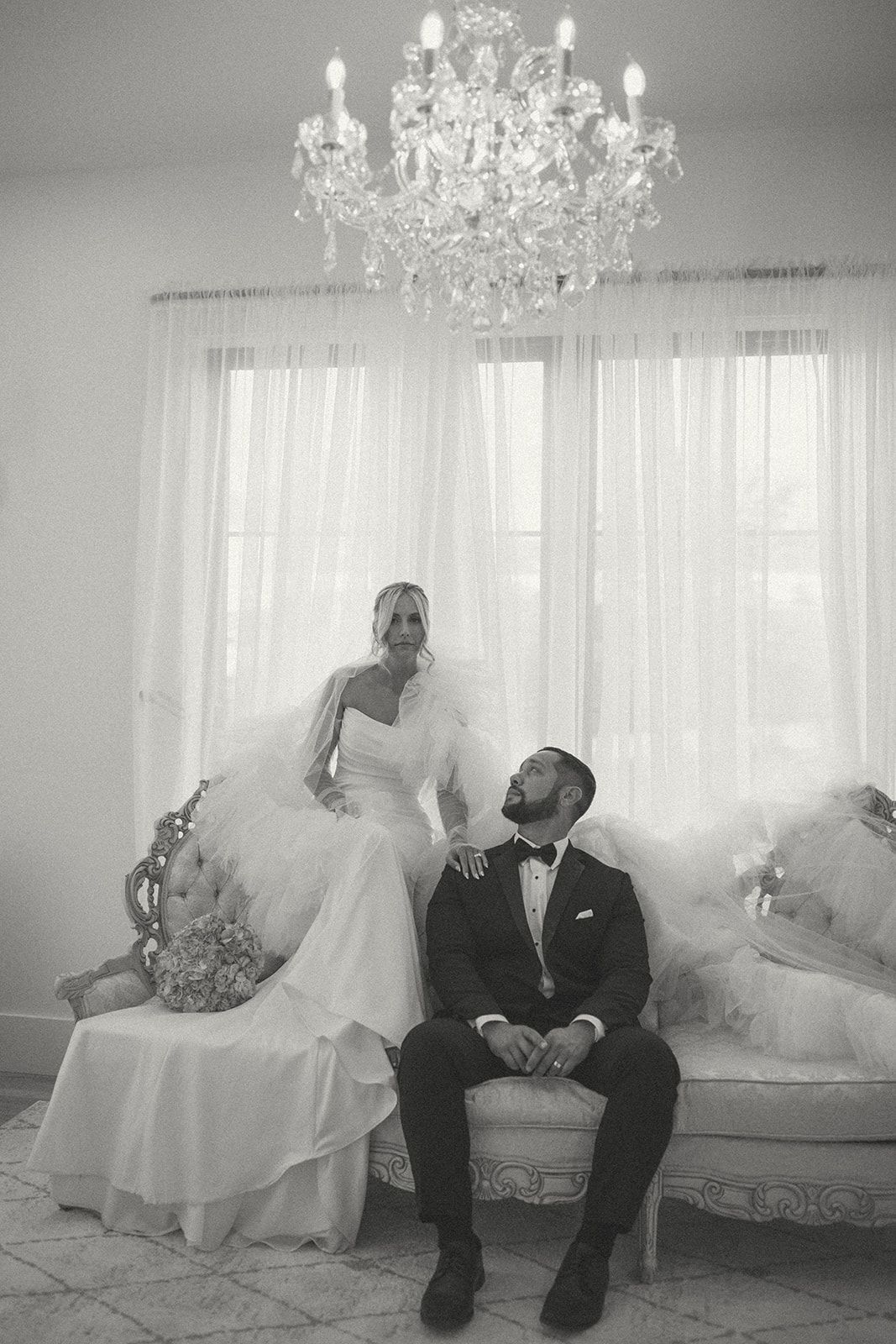 Bride and groom seated in a bright living room beneath a chandelier and window curtains