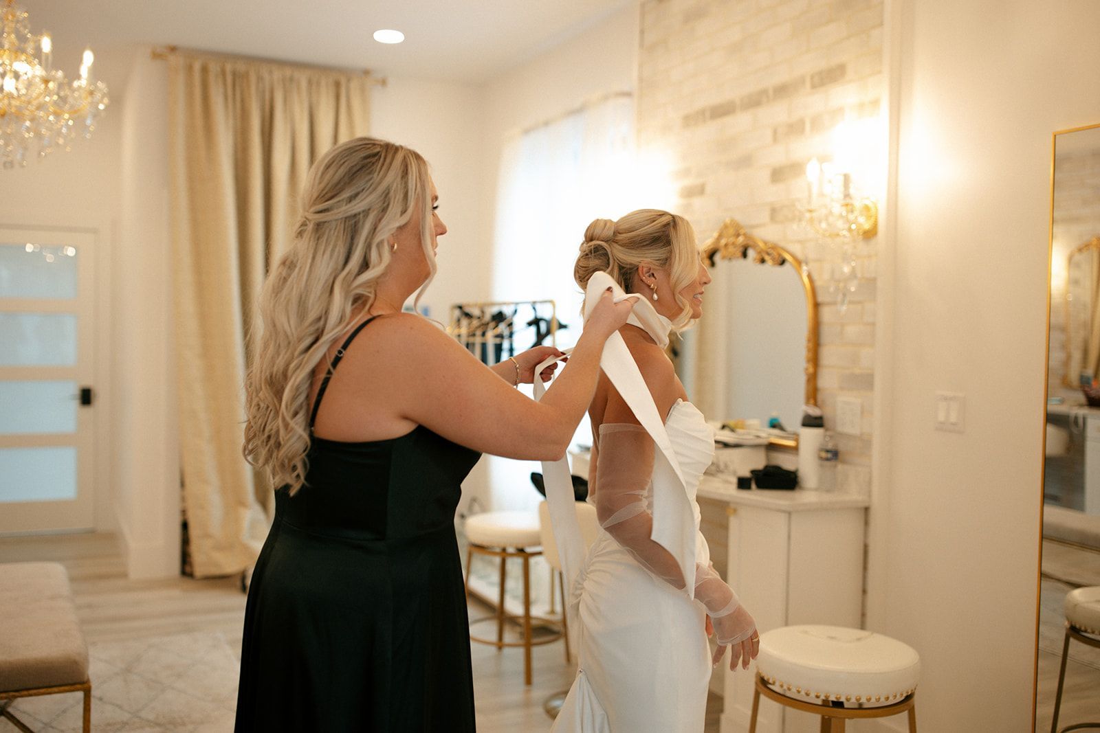 Woman adjusting another woman's dress in a bright fitting room with mirrors and stools