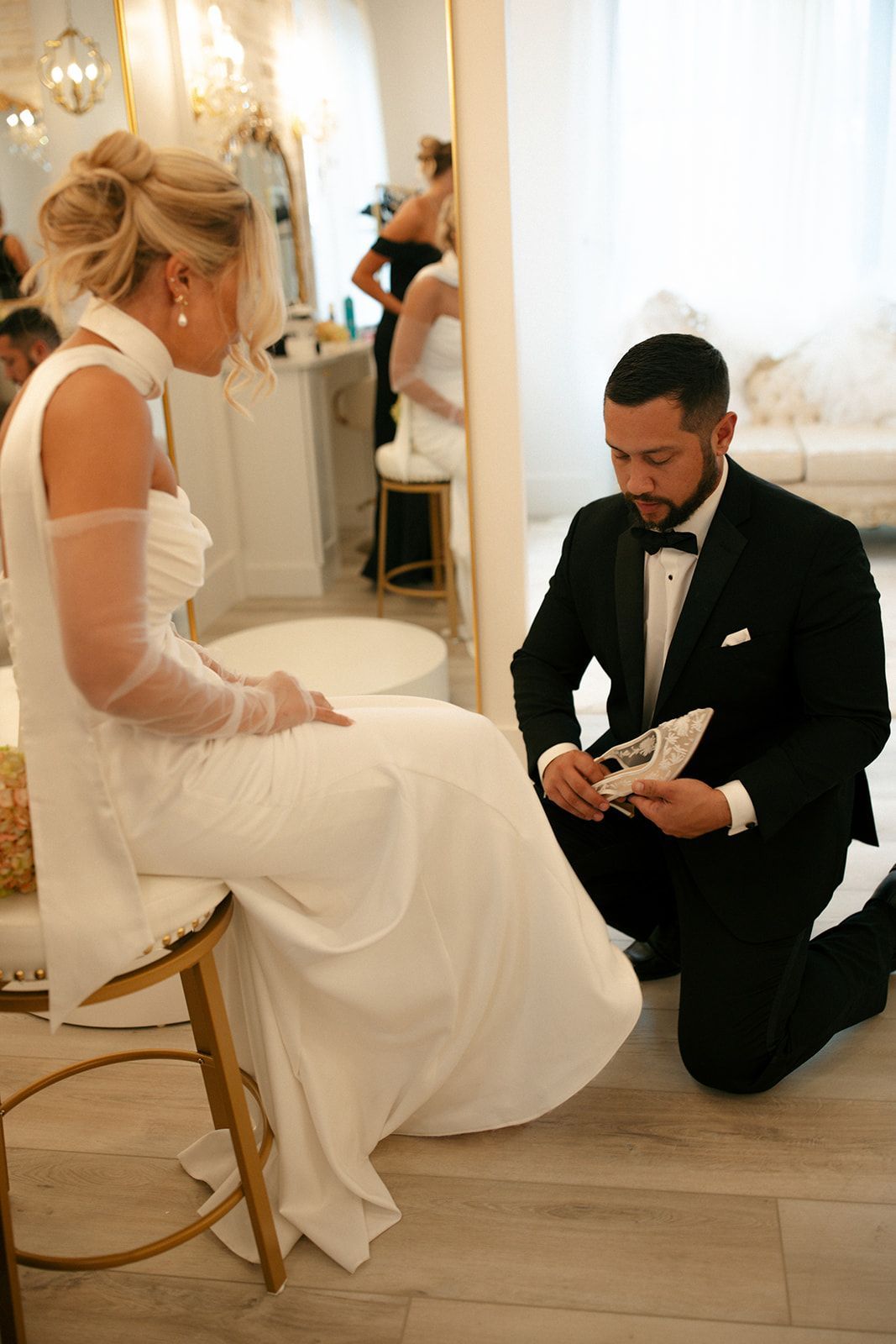 Bride seated in white gown as groom kneels, adjusting her shoe in a bright wedding room