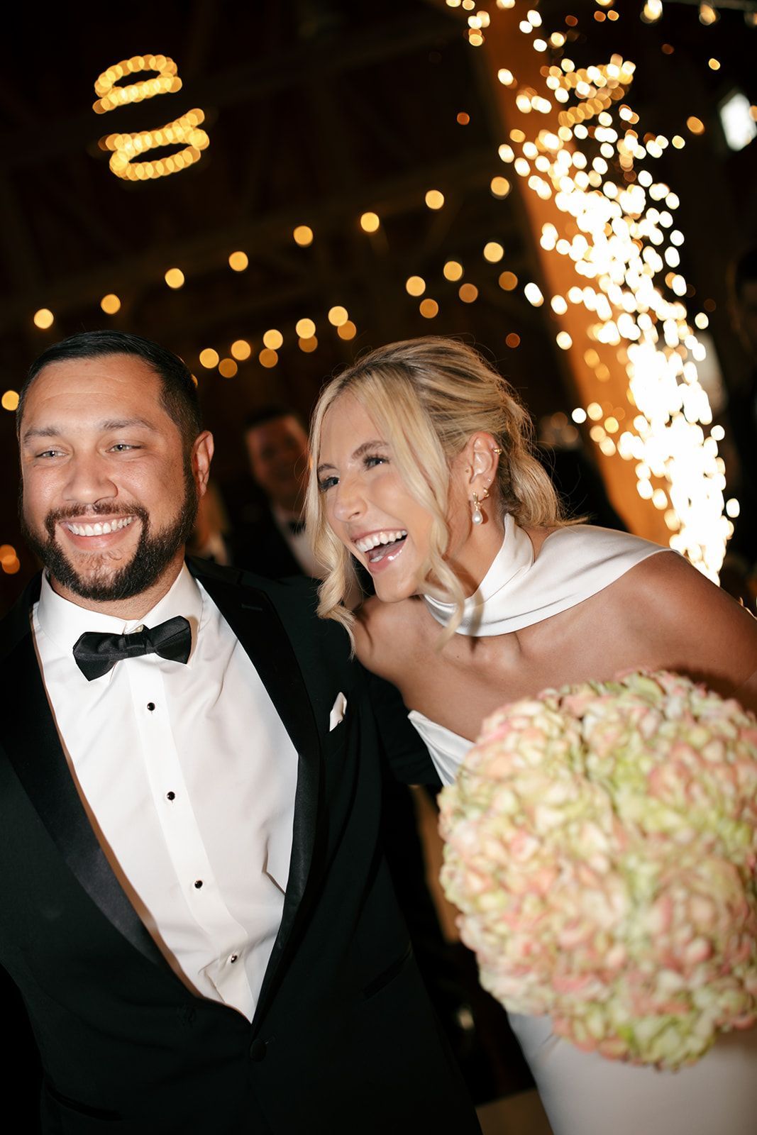 Smiling couple in formalwear at a celebration, with fireworks and a bouquet in the foreground