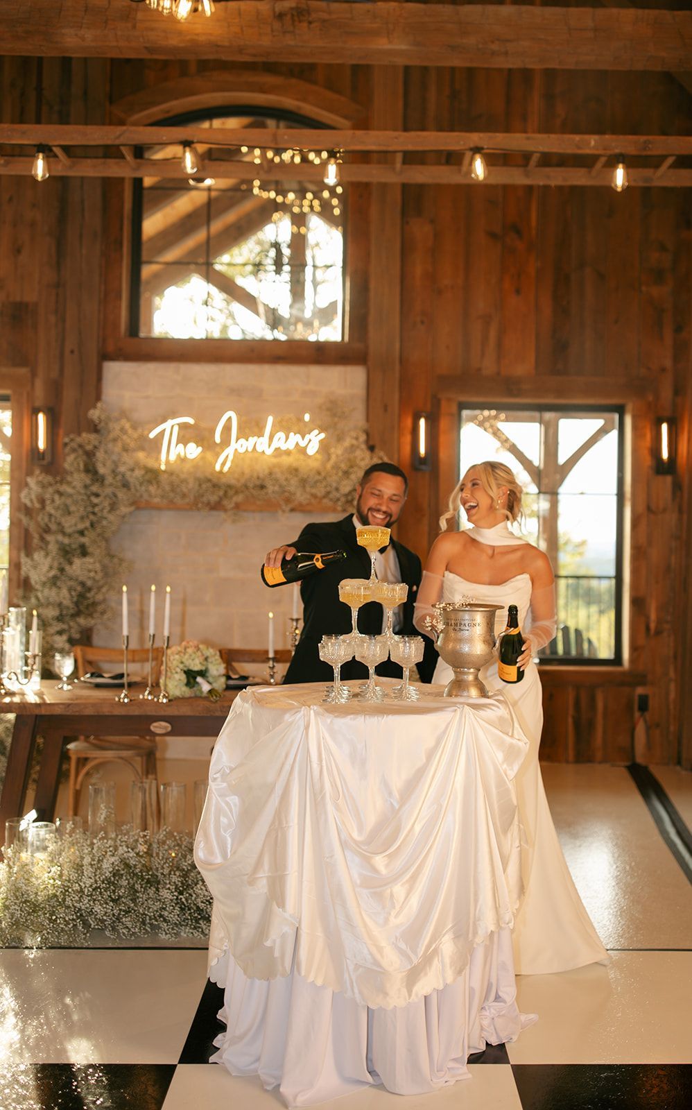 Two people at a champagne tower in a warm-lit rustic reception hall