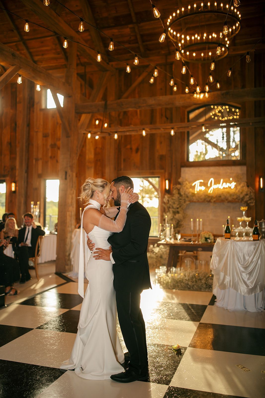 Bride and groom dancing in a rustic barn reception with chandeliers and a black-and-white checkered floor