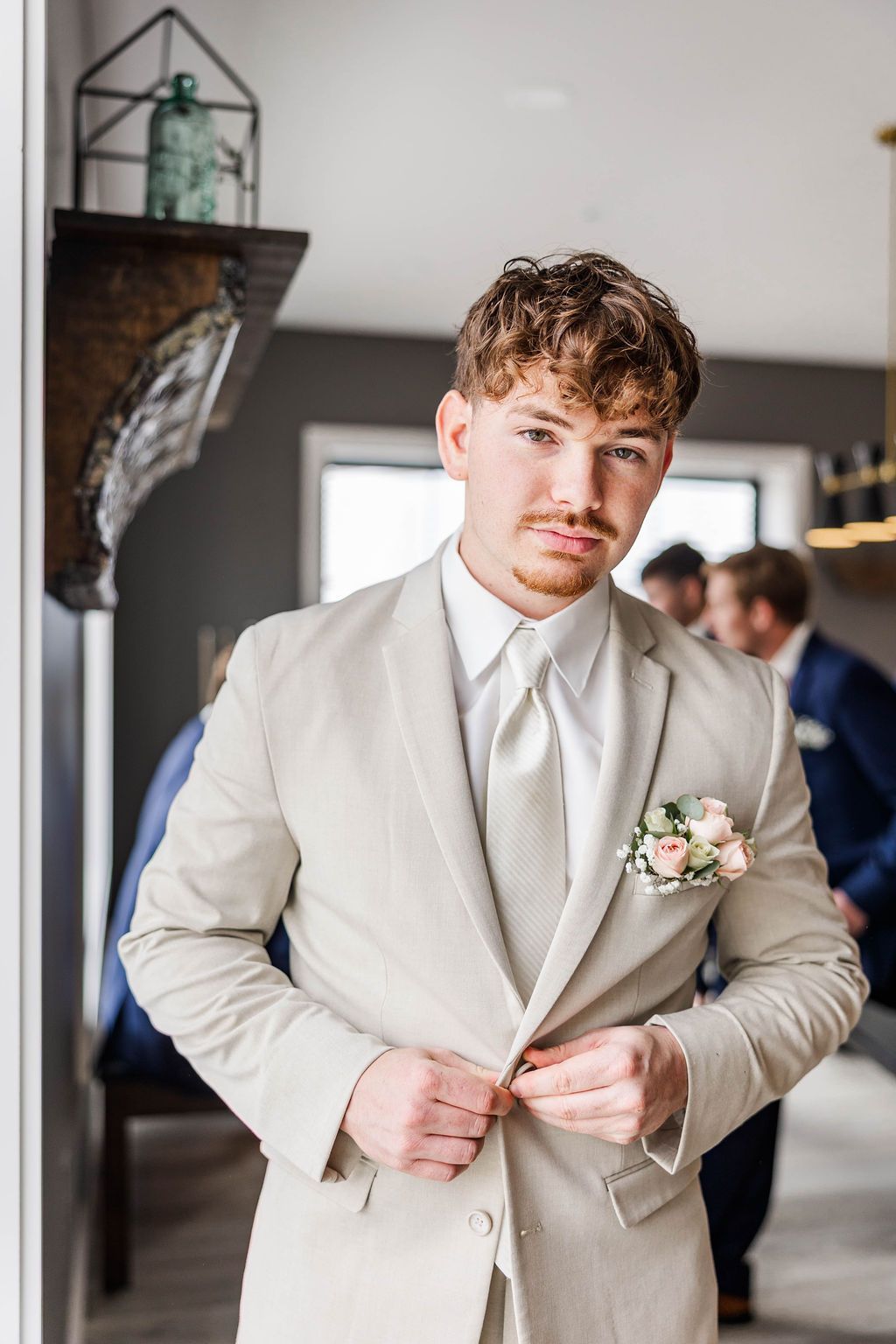 Man in a light beige suit adjusting his jacket indoors, with a boutonniere on his lapel.