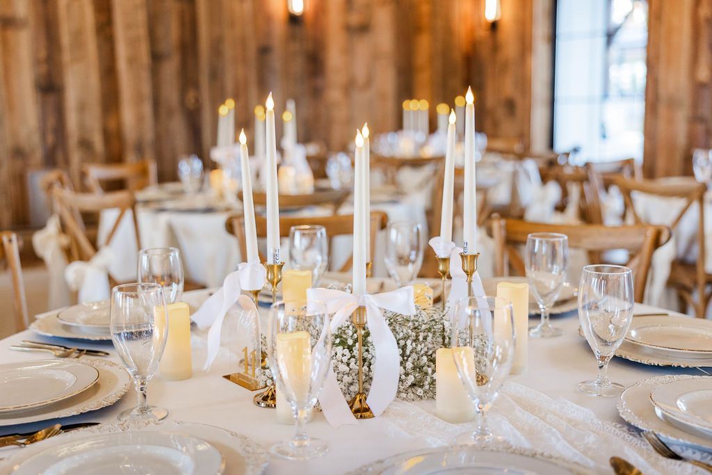 Elegant wedding reception table with white linens, candles, glassware, and gold accents in a rustic venue