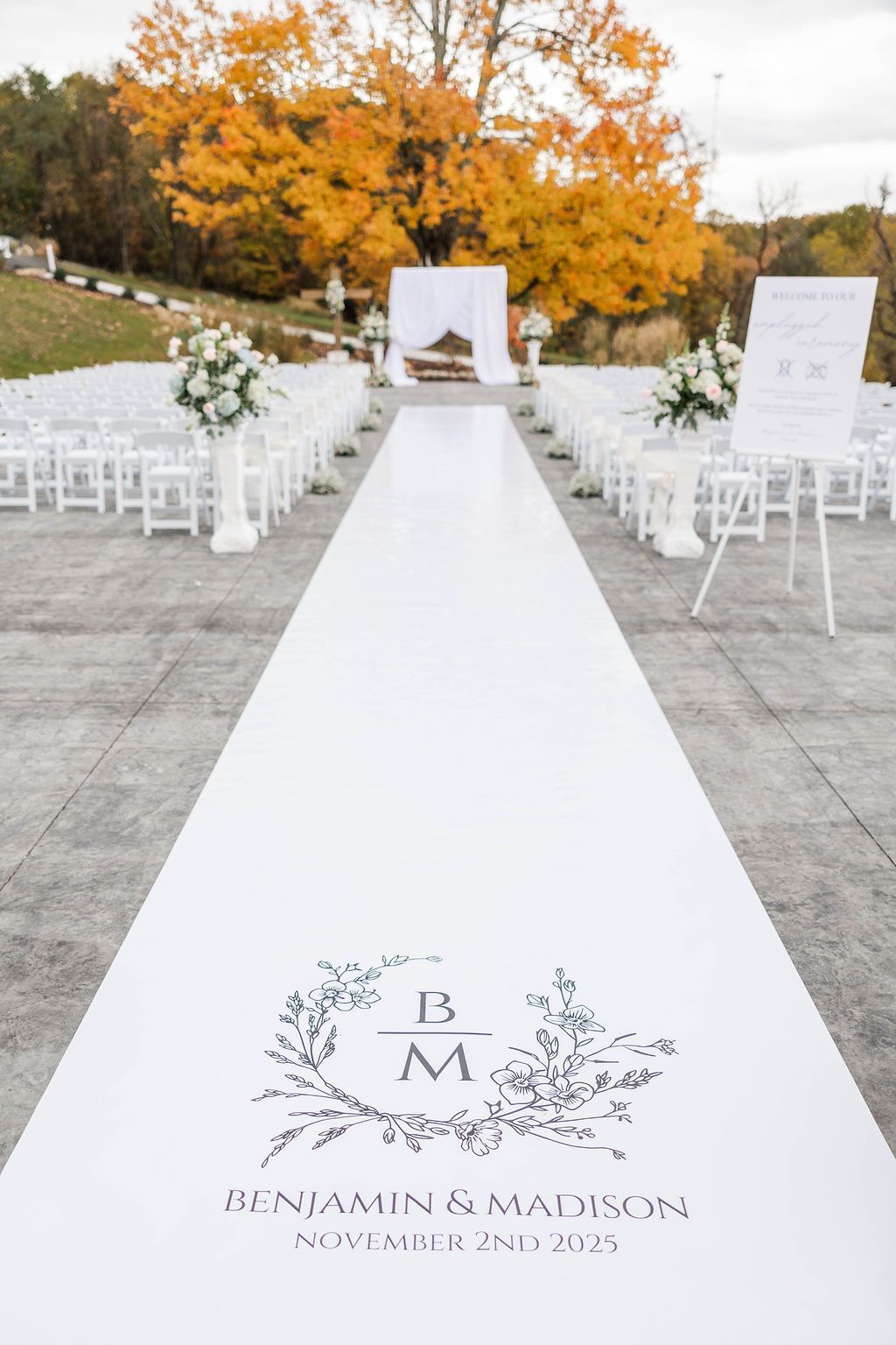 White wedding aisle with chairs and floral arrangements under autumn trees