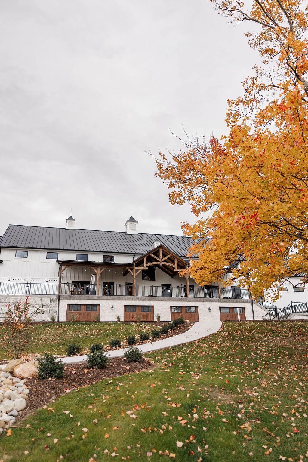 Large white house with a gray roof beside an autumn tree and curving path, with fallen orange leaves.