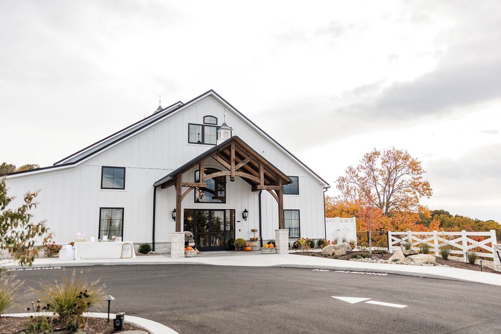 White barn-style building with a covered entrance, gravel driveway, and autumn trees in front