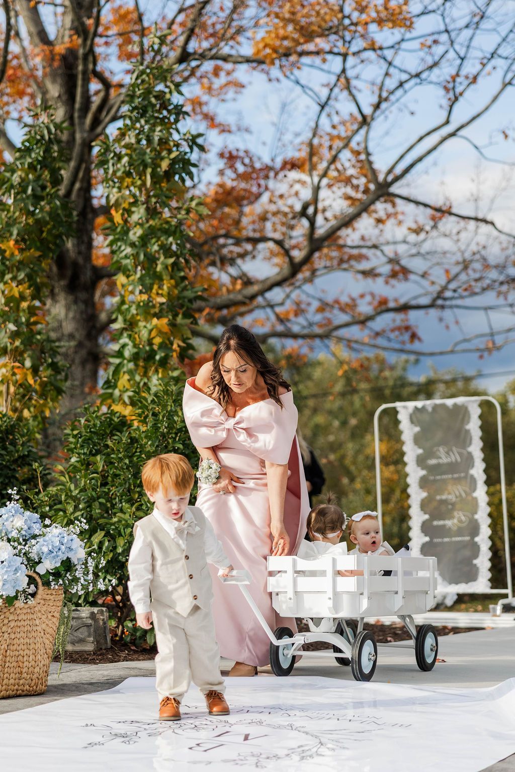 Woman and child in pastel outfits outdoors beside a white cart under autumn trees