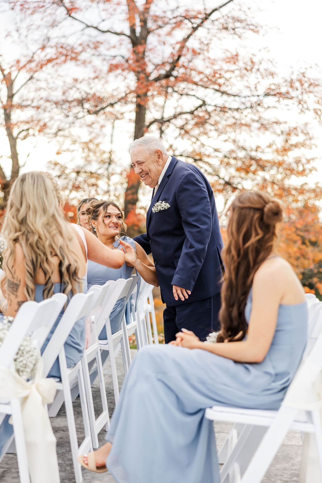 Outdoor wedding ceremony with guests seated on white chairs, greeting an older man at the aisle