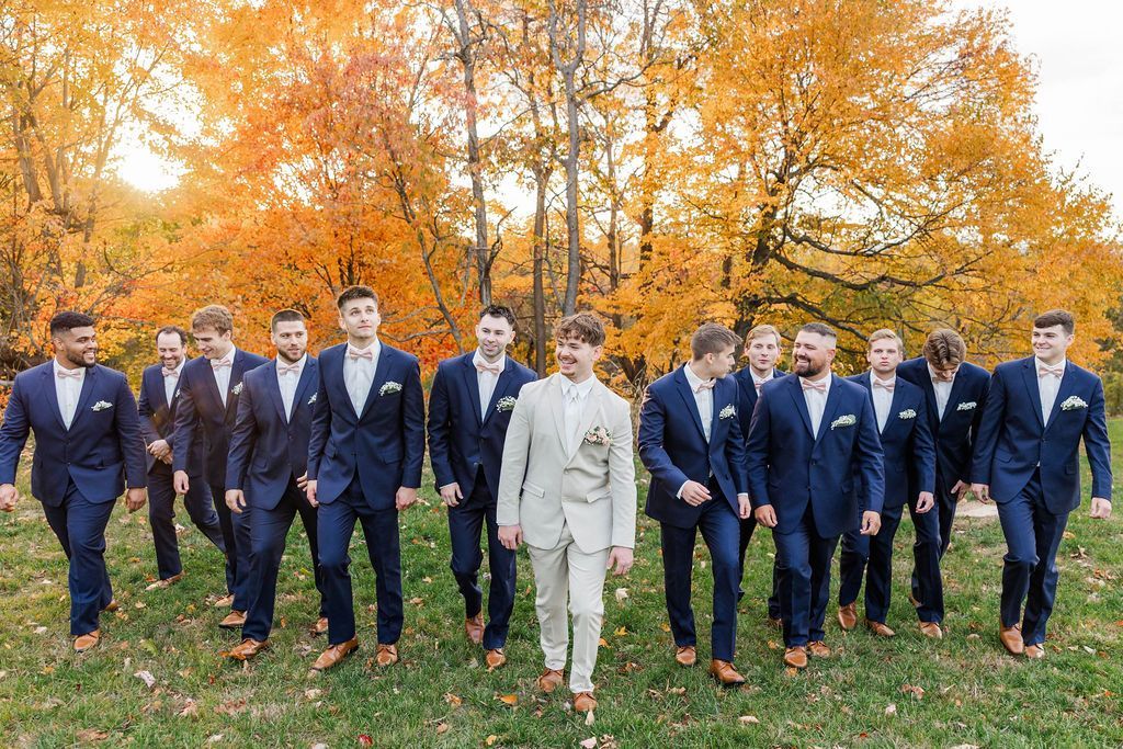 Group of men in suits walking outdoors on grass in front of bright autumn trees