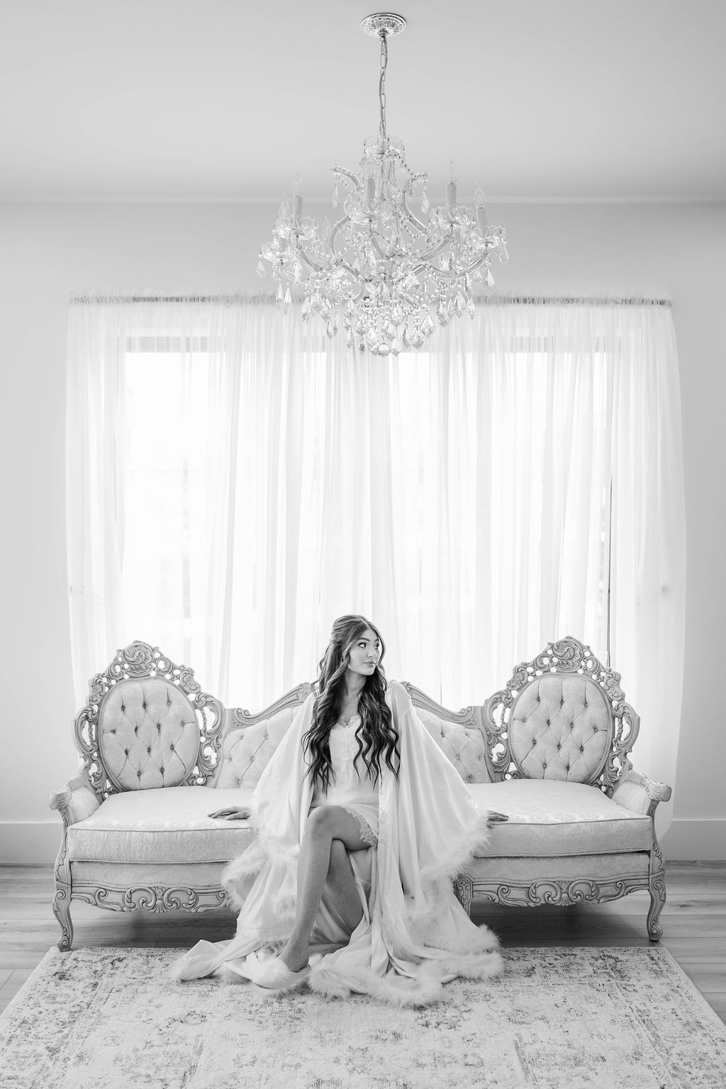 Woman seated on a tufted sofa in a bright room with sheer curtains and a chandelier.