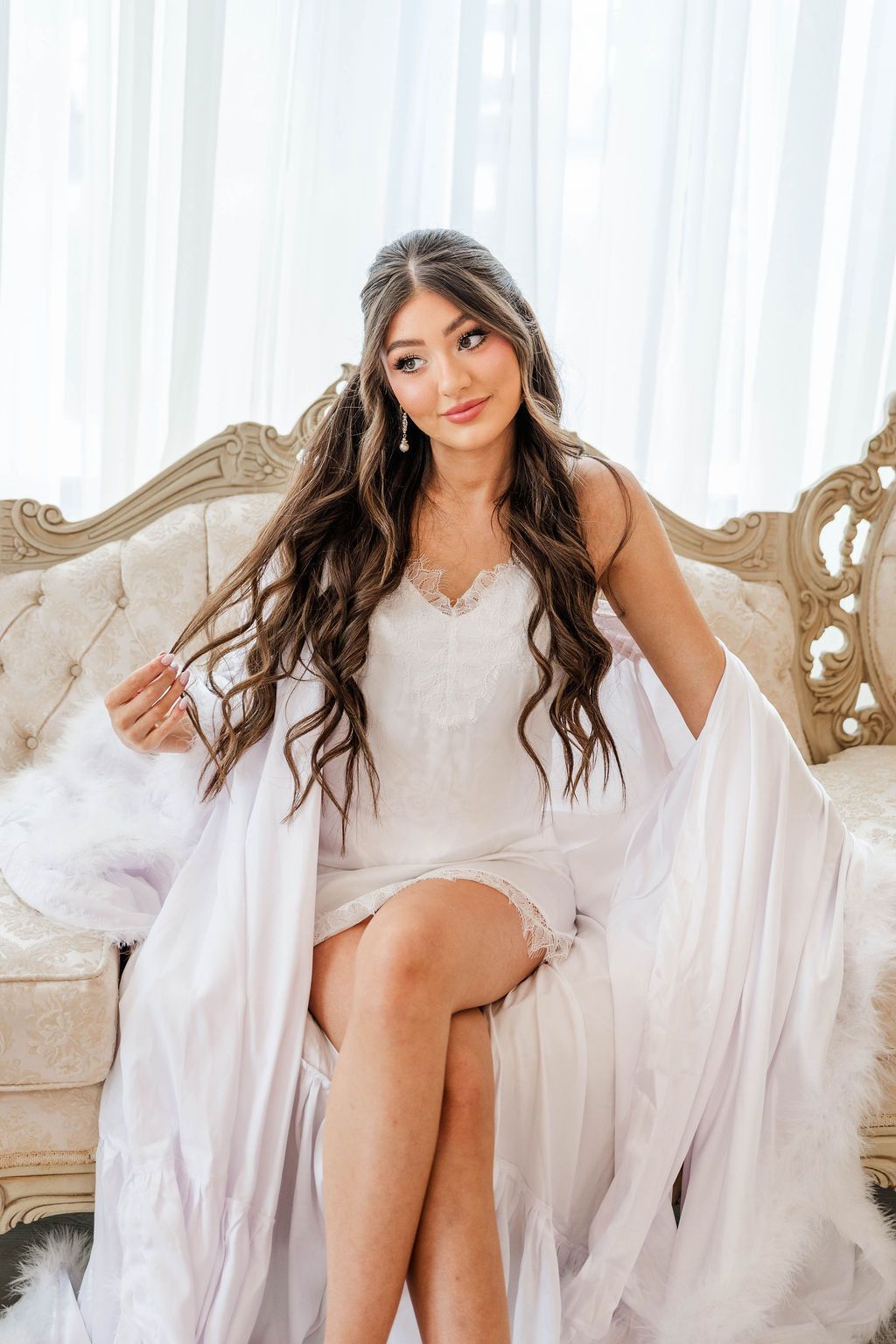 Woman in a white dress seated on an ornate sofa in a bright room with sheer curtains.