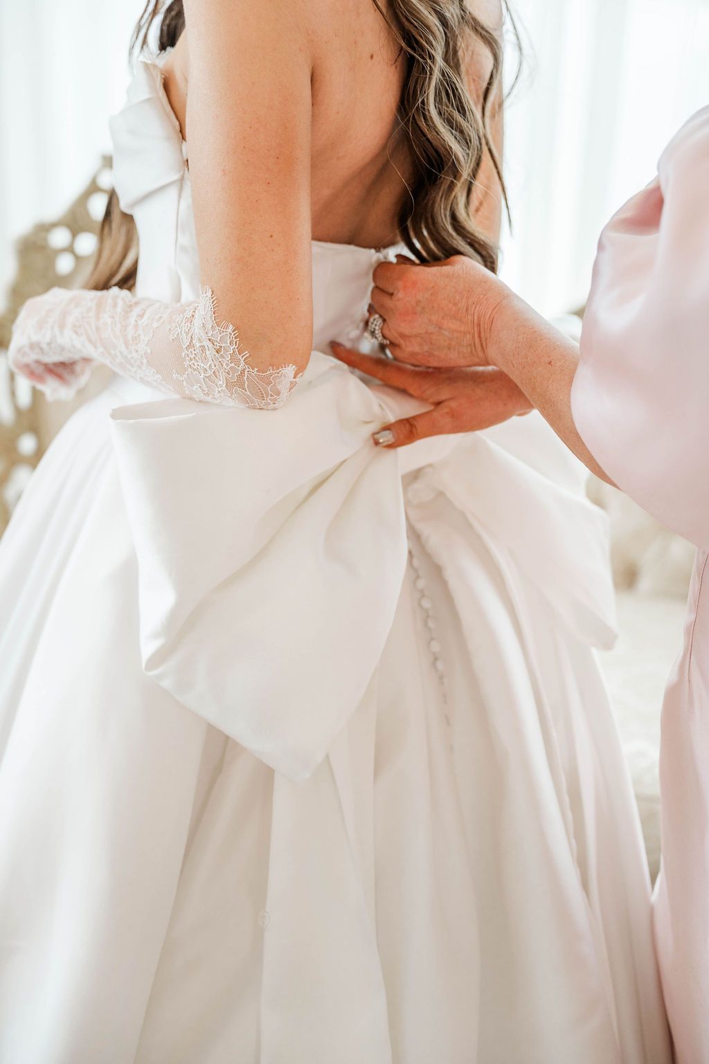 Hands fastening the back of a white wedding dress with lace sleeves and a large bow.