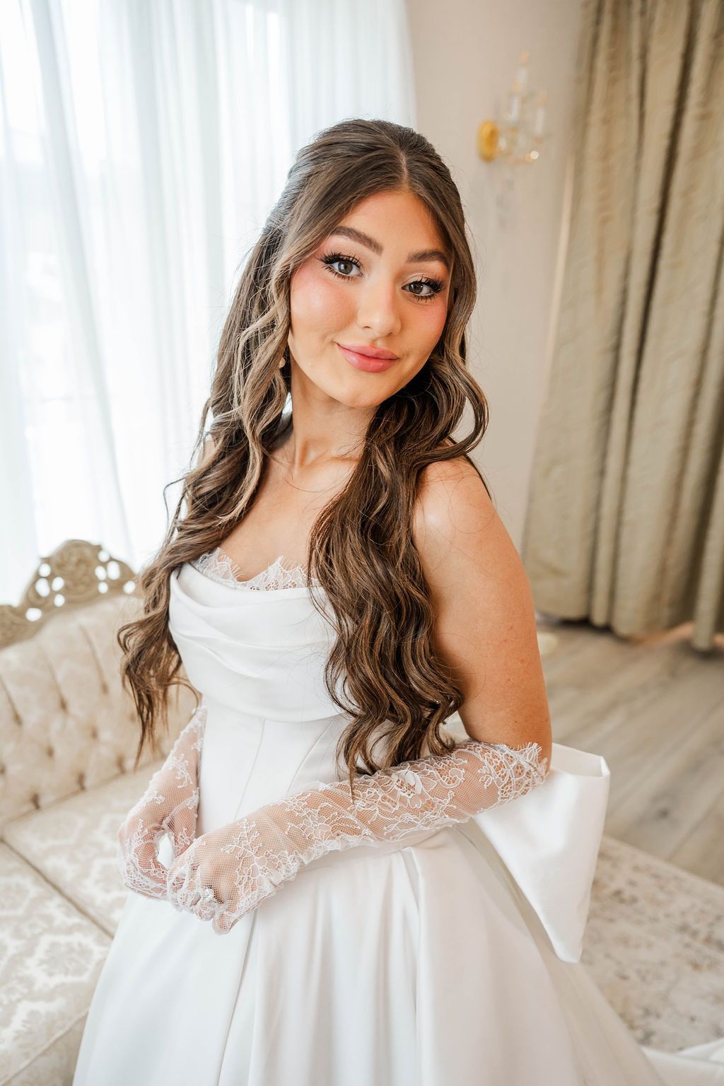 Bride in a white wedding dress posing indoors by a window, with long wavy hair and lace gloves
