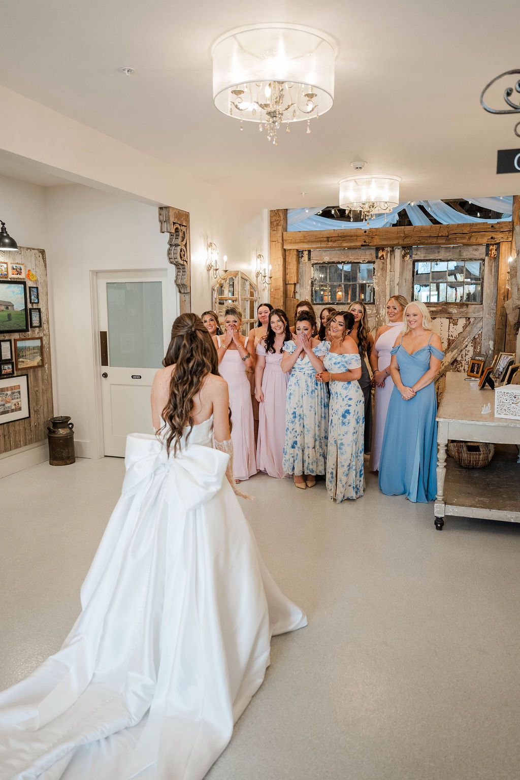 Bride in white gown faces bridesmaids in pastel dresses inside a bright room