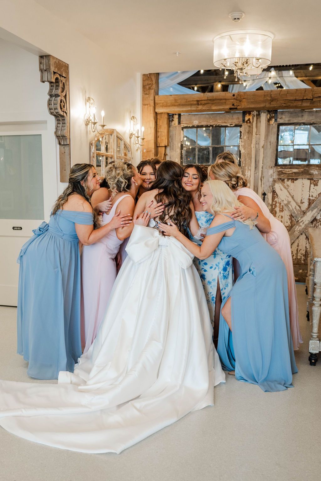 Bridal party in blue dresses hugs a bride in a white gown inside a rustic venue.
