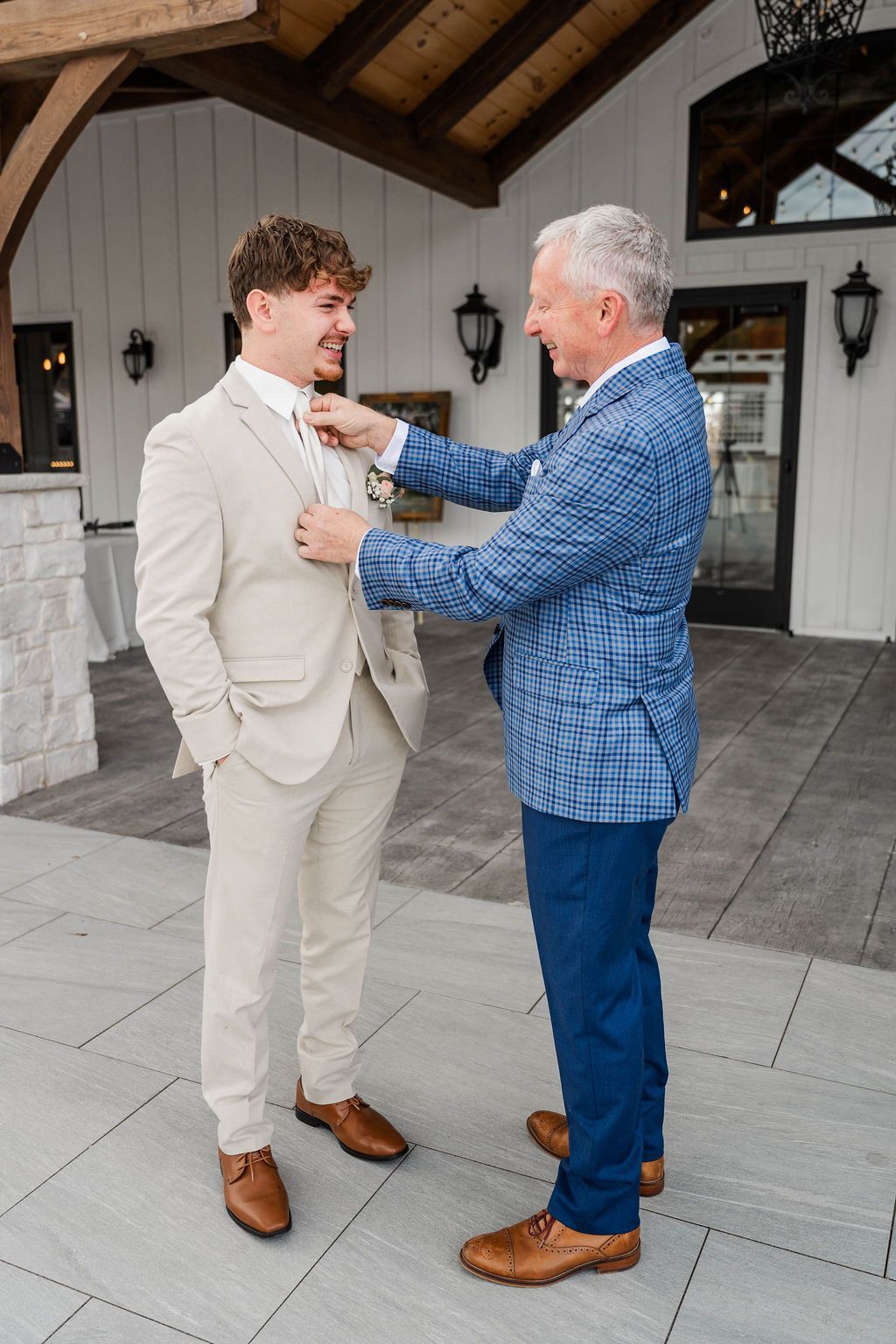 Two men adjust a boutonniere on a cream suit outdoors by a white building.