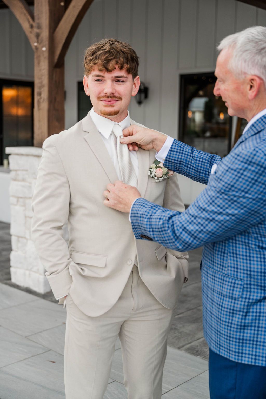 Man adjusting a boutonniere on another man’s beige suit outside a venue
