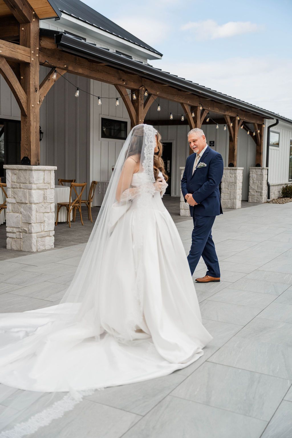 Bride in a white gown walks toward a man in a navy suit outside a rustic venue.