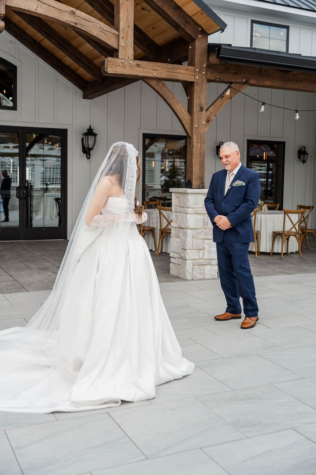 Bride in white gown and veil faces groom in navy suit during outdoor wedding ceremony under a wooden pavilion