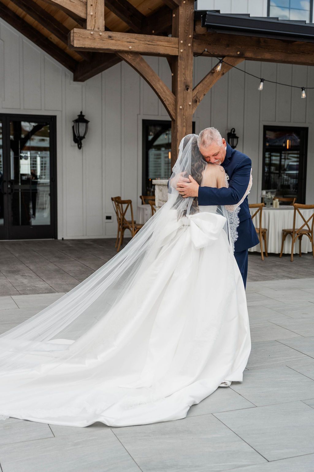 Bride in a long white gown hugs a man in a blue suit outside a rustic building.