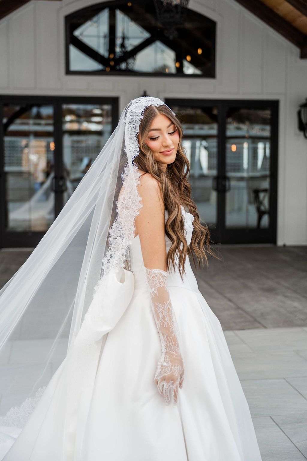 Bride in a white wedding dress and veil standing outside a modern building, smiling back over her shoulder