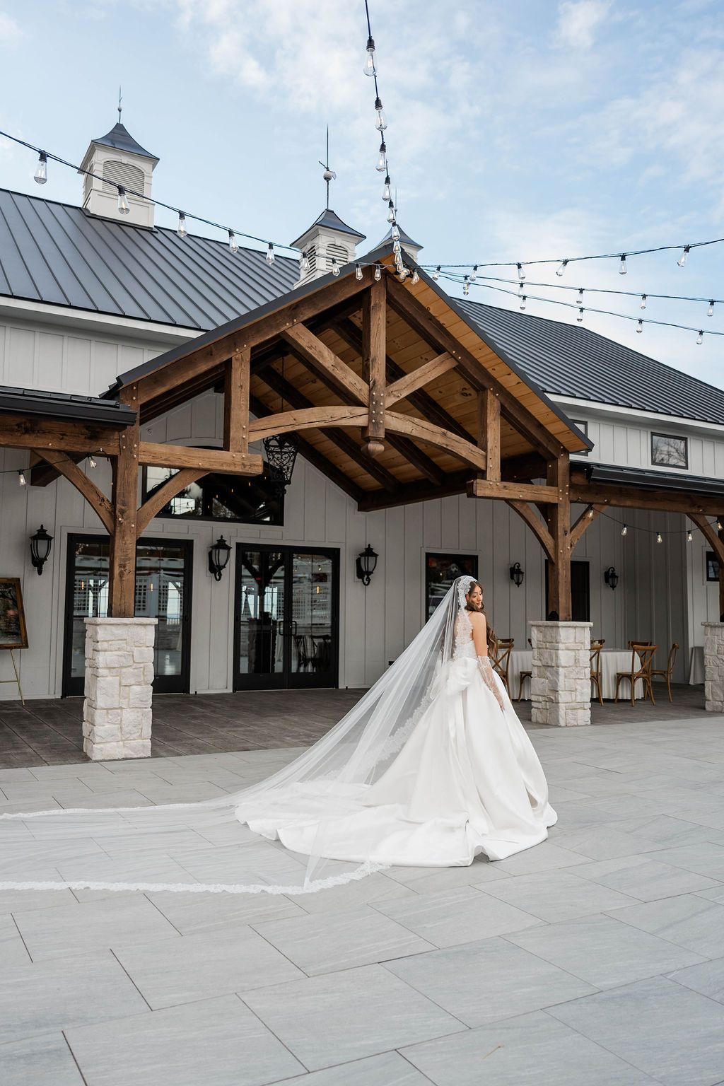 Bride in a white gown and veil standing under a wooden pavilion outside a building