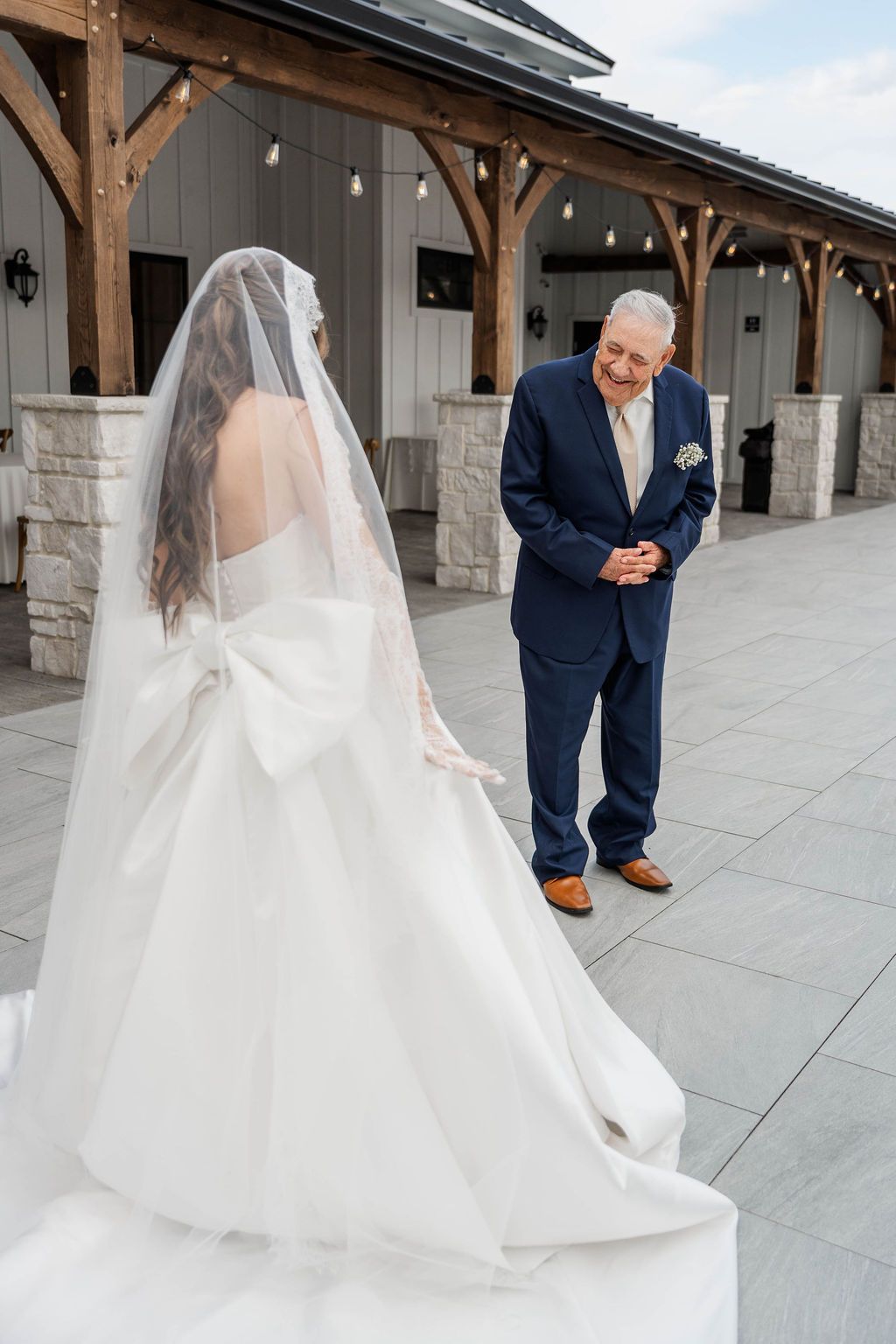 Bride in white veil faces man in navy suit at an outdoor wedding venue
