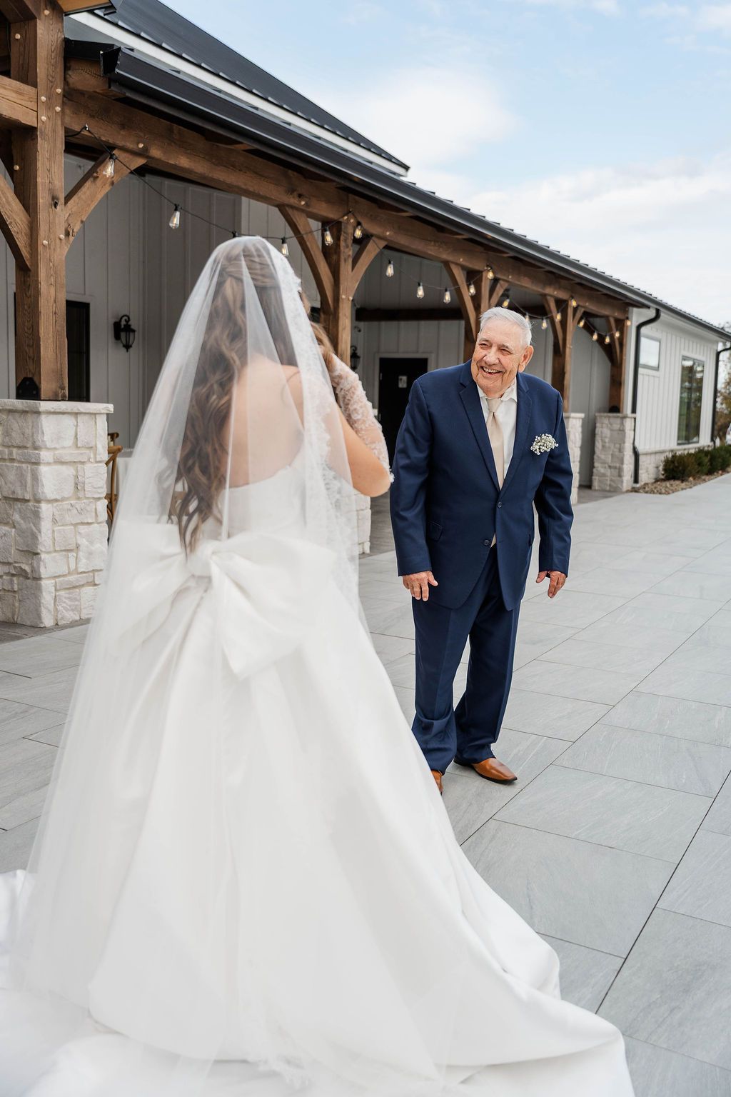 Bride in white veil walks toward smiling man in navy suit beside rustic venue