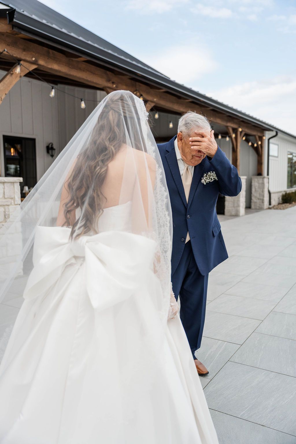 Bride in a white gown and veil walks beside an older man in a blue suit outdoors by a modern building