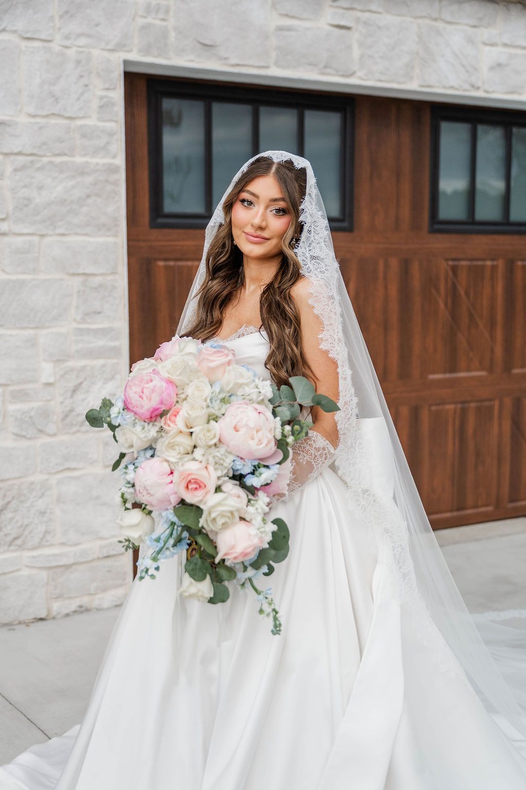 Bride in a white wedding gown holding a pink-and-white bouquet beside a wooden door