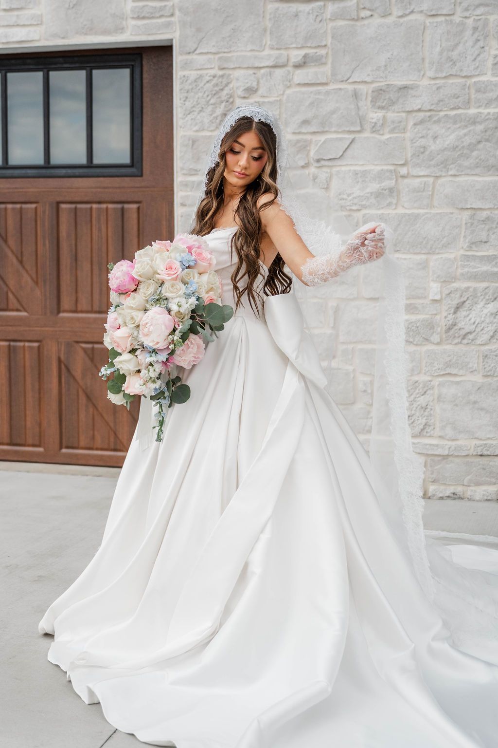 Bride in white gown holding a pastel bouquet beside a stone wall and wooden door