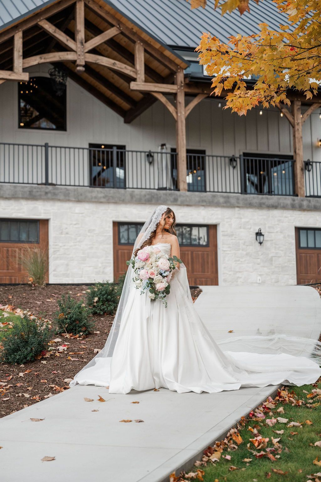 Bride in a white gown holding a bouquet, standing outside a rustic building with autumn leaves.