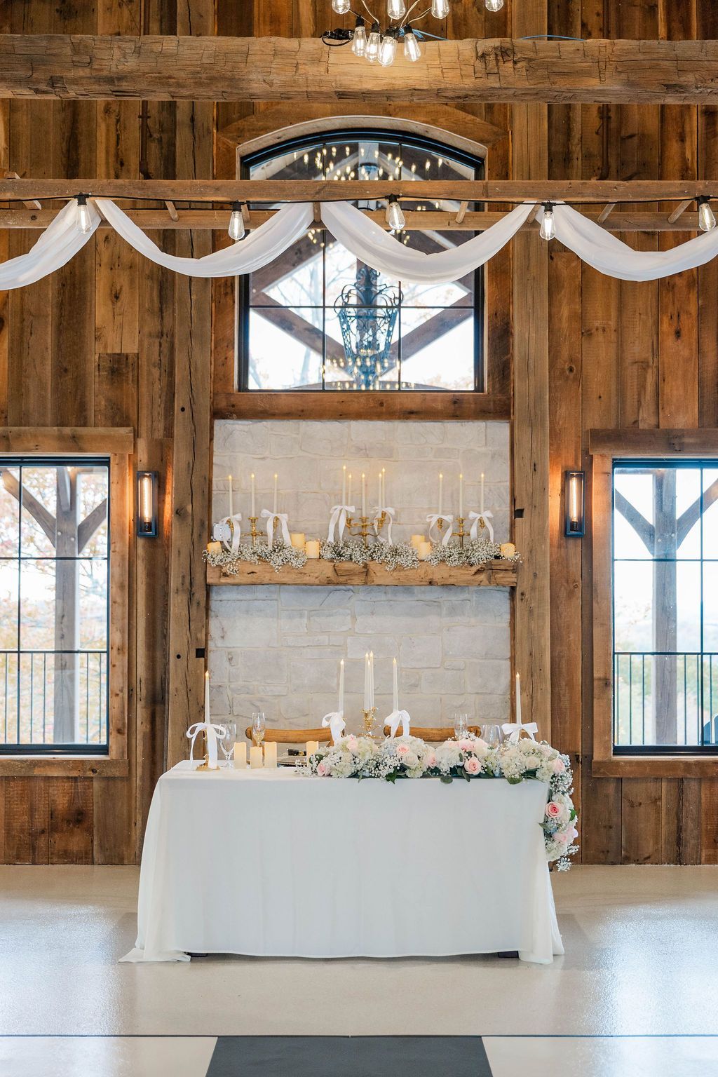 Rustic wedding reception table with white linens, floral decor, and soft draped fabric in a wood barn setting