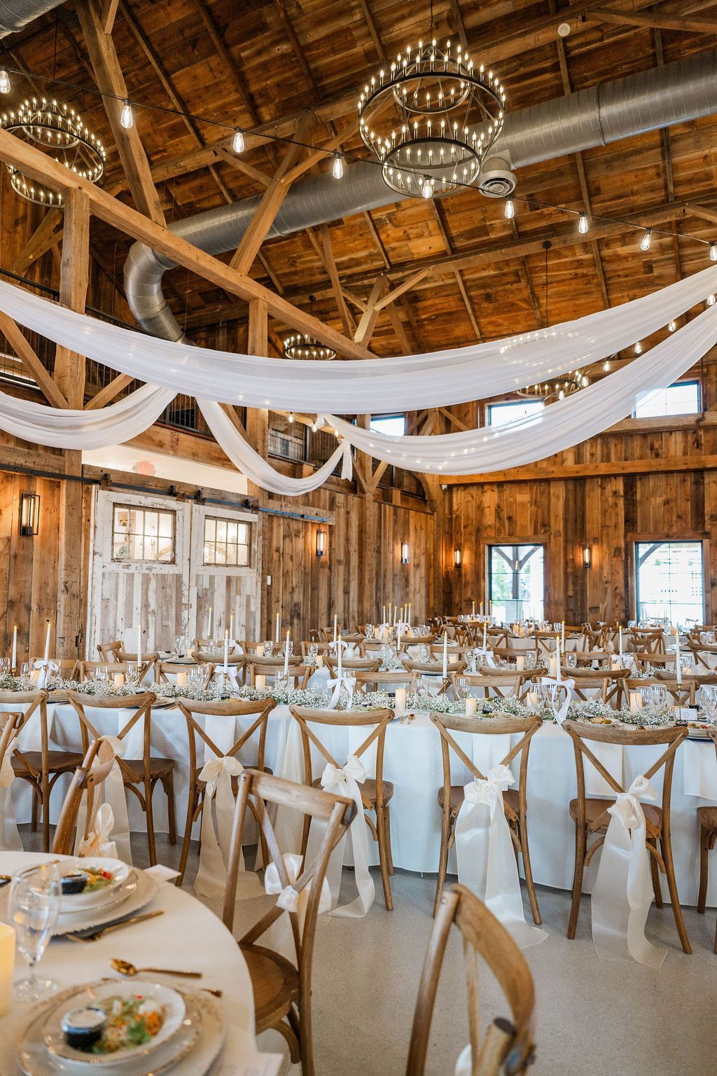 Rustic barn wedding reception with white draped fabric, chandeliers, and rows of decorated tables