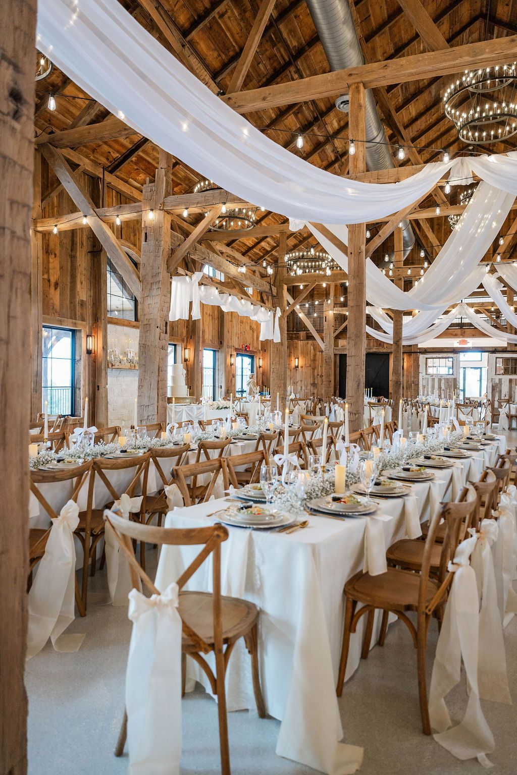 Rustic barn wedding reception with long white-draped tables, wooden chairs, and sheer fabric hanging from the rafters