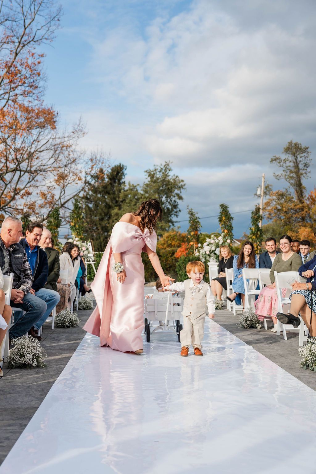 Outdoor wedding aisle with a bride in pink and a child walking ahead on a white runner, guests seated on both sides