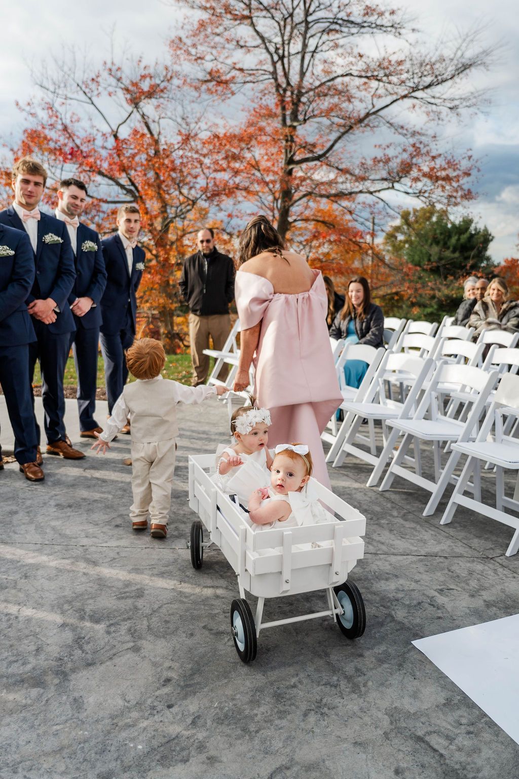 Wedding guests stand outdoors; a woman pulls a white wagon with a baby doll beside white chairs and autumn trees.