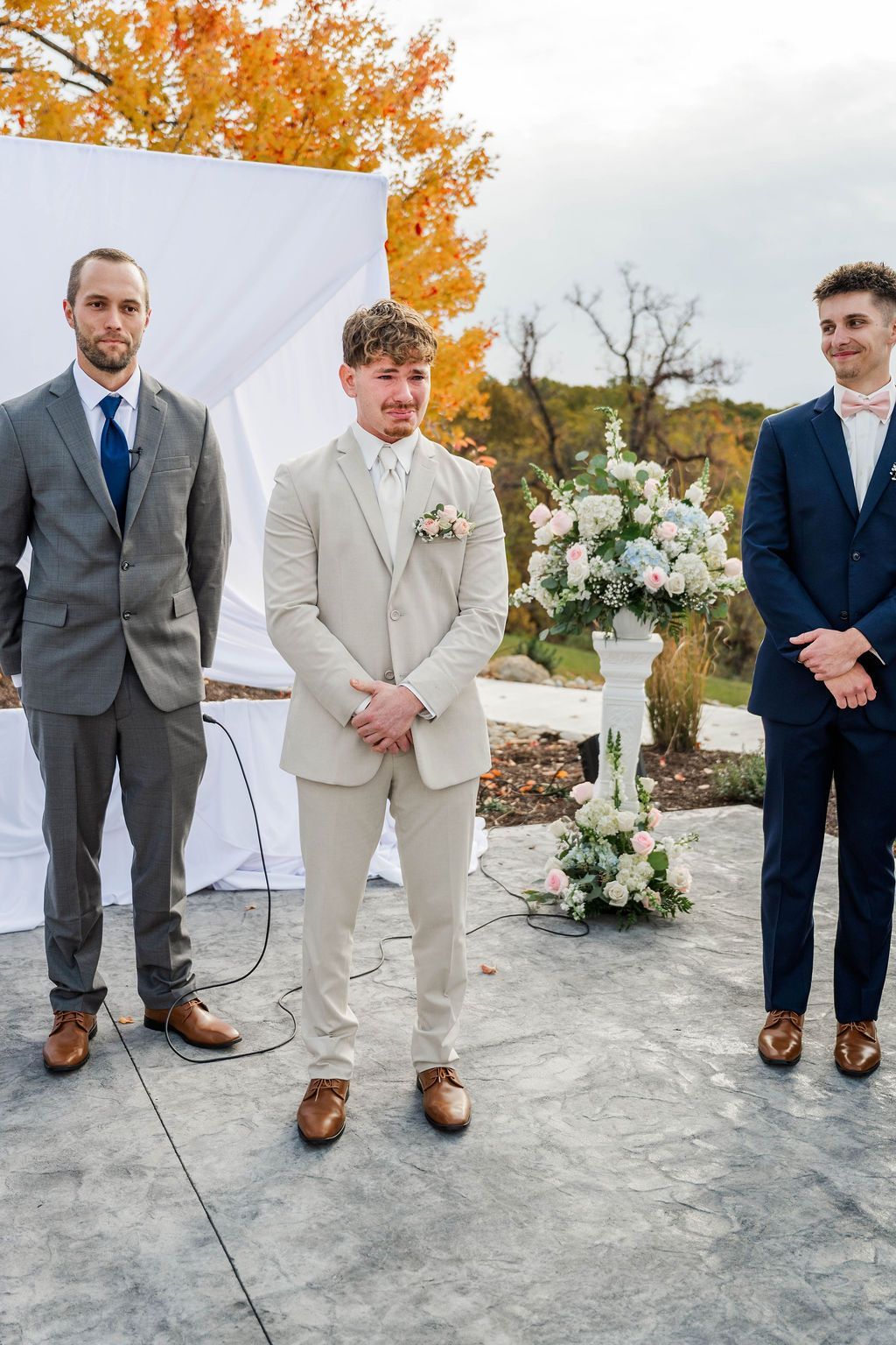 Three men in suits standing outdoors at a decorated wedding ceremony, with autumn trees and floral arrangements.