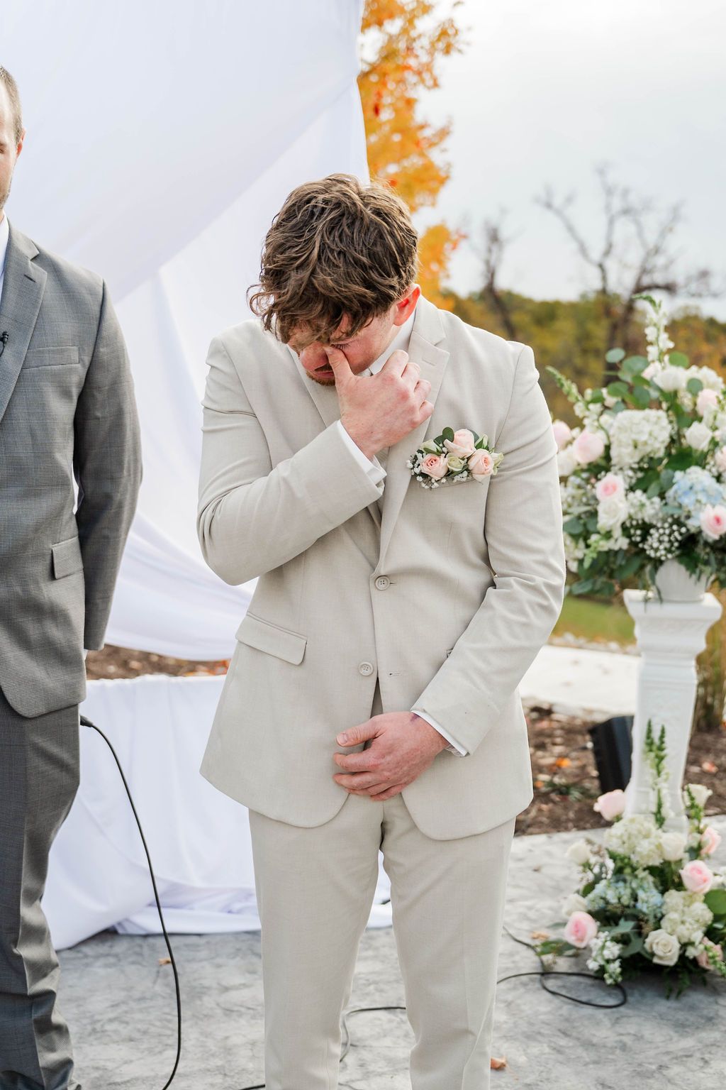 Person in a beige suit covers their face at an outdoor wedding beside floral arrangements.