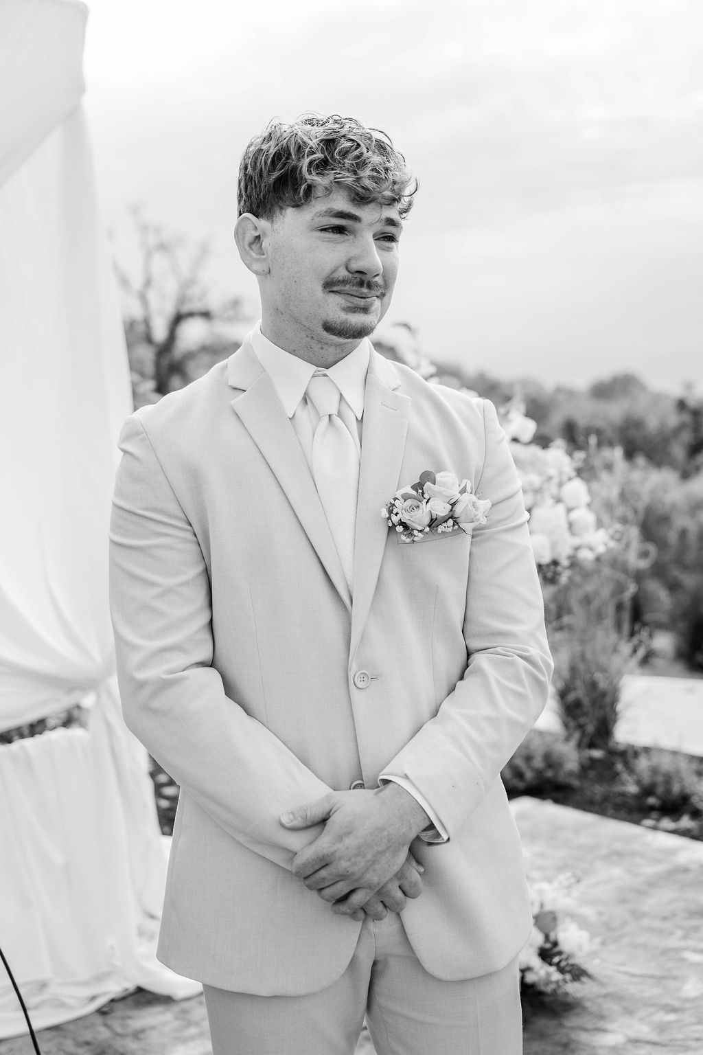 Man in a light suit standing outdoors with hands clasped, wearing a boutonniere.